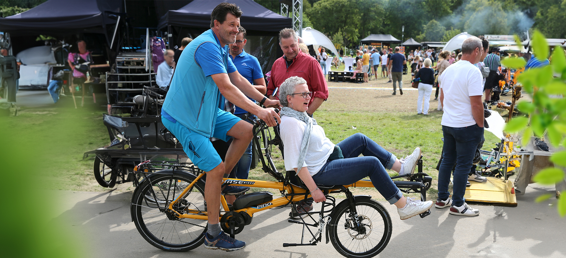 Eine Frau sitzt mit einem Mann auf einem Fahrrad, das einem Tandem ähnlich ist. Beide fahren durch den Wiehlpark, im Hintergrund sind viele Menschen zu sehen.