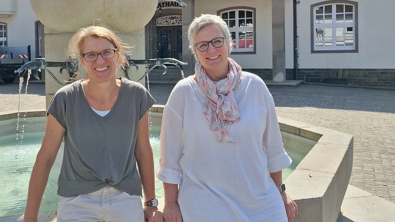 Zwei Frauen mit Brillen stehen an einem sonnigen Tag vor einem Steinbrunnen auf einem Platz im Freien und lächeln.