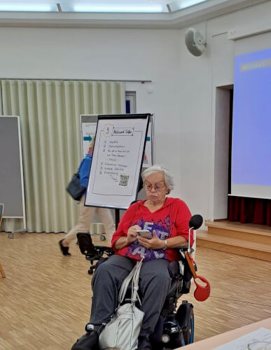 Eine ältere Frau in einem motorisierten Rollstuhl benutzt ihr Smartphone in einem Besprechungsraum mit einem Präsentationsbildschirm und einem Flipchart im Hintergrund.
