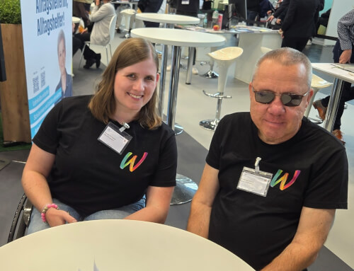 Zwei Personen, die passende schwarze T-Shirts mit einem bunten W tragen, sitzen an einem runden Tisch in einem Veranstaltungsraum mit anderen Teilnehmern im Hintergrund.