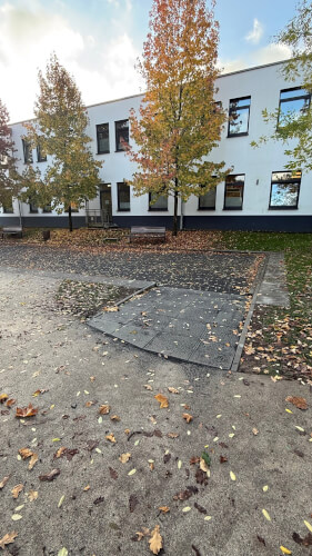 Eine Rollstuhlrampe führt zu einem erhöhten Weg vor einem weißen Gebäude mit großen Fenstern und Herbstbäumen. Gefallene Blätter bedecken den Boden.