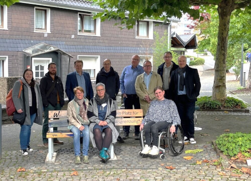 Eine Gruppe von elf Erwachsenen, darunter eine Person im Rollstuhl, posiert im Freien neben einer Bank und einem Schild auf einem Kopfsteinpflaster vor Wohnhäusern.