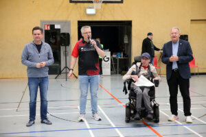 Vier Männer stehen auf dem Boden einer Sporthalle; einer sitzt im Rollstuhl, einer spricht in ein Mikrofon, zwei weitere stehen auf beiden Seiten, alle mit Blick in die Kamera.