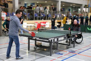 Zwei Männer, einer davon im Rollstuhl, spielen Tischtennis in einer Sporthalle, während im Hintergrund Zuschauer und andere Personen zu sehen sind.