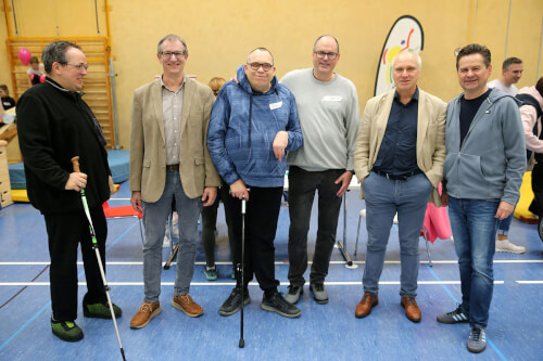 Sechs Männer stehen in einer Turnhalle zusammen, einige mit Stöcken, und posieren für ein Gruppenfoto. Im Hintergrund sind Stühle, Turngeräte und andere Personen zu sehen.