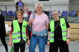 Drei Erwachsene stehen in einem Haus, zwei mit neongrünen Warnwesten und einer in Freizeitkleidung. Sie posieren und lächeln in die Kamera. Im Hintergrund sind blaue Sitze und Menschen zu sehen.
