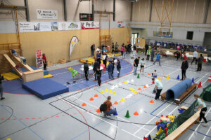 In einer Turnhalle werden verschiedene körperliche Aktivitäten und Hindernisläufe durchgeführt. Geräte und farbige Kegel sind auf dem Boden aufgestellt. An der Wand hängen Schilder und Transparente.