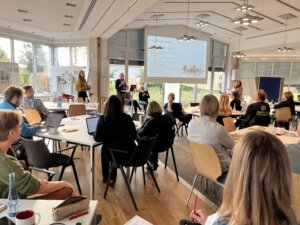 Die Teilnehmer eines Workshops oder Seminars sitzen an Tischen in einem Konferenzraum und hören den Referenten zu, die ihre Folien im vorderen Teil des Raumes präsentieren.