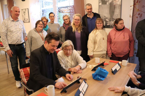Eine Gruppe von Menschen, darunter Senioren und Erwachsene, versammelt sich in einem Innenraum um einen Tisch und beobachtet einen Mann, der ein Tablet benutzt, während andere ihn beobachten und lächeln.