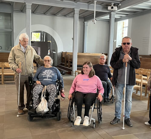 Fünf Erwachsene, zwei im Rollstuhl und zwei mit weißen Stöcken, stehen und sitzen zusammen in einem geräumigen, gut beleuchteten Raum mit Holzstühlen und hohen Decken.