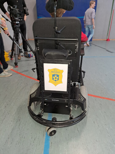 Ein Elektrorollstuhl mit einem BSV Bielstein-Schriftzug auf der Rückseite ist in einer Turnhalle geparkt, im Hintergrund sind Menschen und eine Kamera zu sehen.