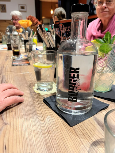 Eine durchsichtige, mit Wasser gefüllte Flasche mit der Aufschrift "Burger Queen" steht auf einem hölzernen Restauranttisch inmitten von Getränken und Gläsern, um die herum Menschen sitzen.