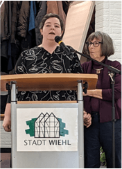 Zwei Frauen stehen an einem Podium mit einem Mikrofon; eine spricht, während die andere ihre Hand hält. Auf dem Podium steht ein Schild der Stadt Wiehl.