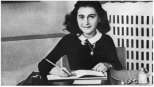 Ein junges Mädchen mit dunklem Haar sitzt an einem Schreibtisch, schreibt in ein Notizbuch und lächelt in die Kamera. Es scheint sich um einen Innenraum zu handeln, in dem Schreibzeug auf dem Schreibtisch liegt.