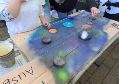 Man sprüht ein Weltraummotiv mit Planeten und Linien auf ein Holzbrett und verwendet Tassen und Deckel als Schablonen.