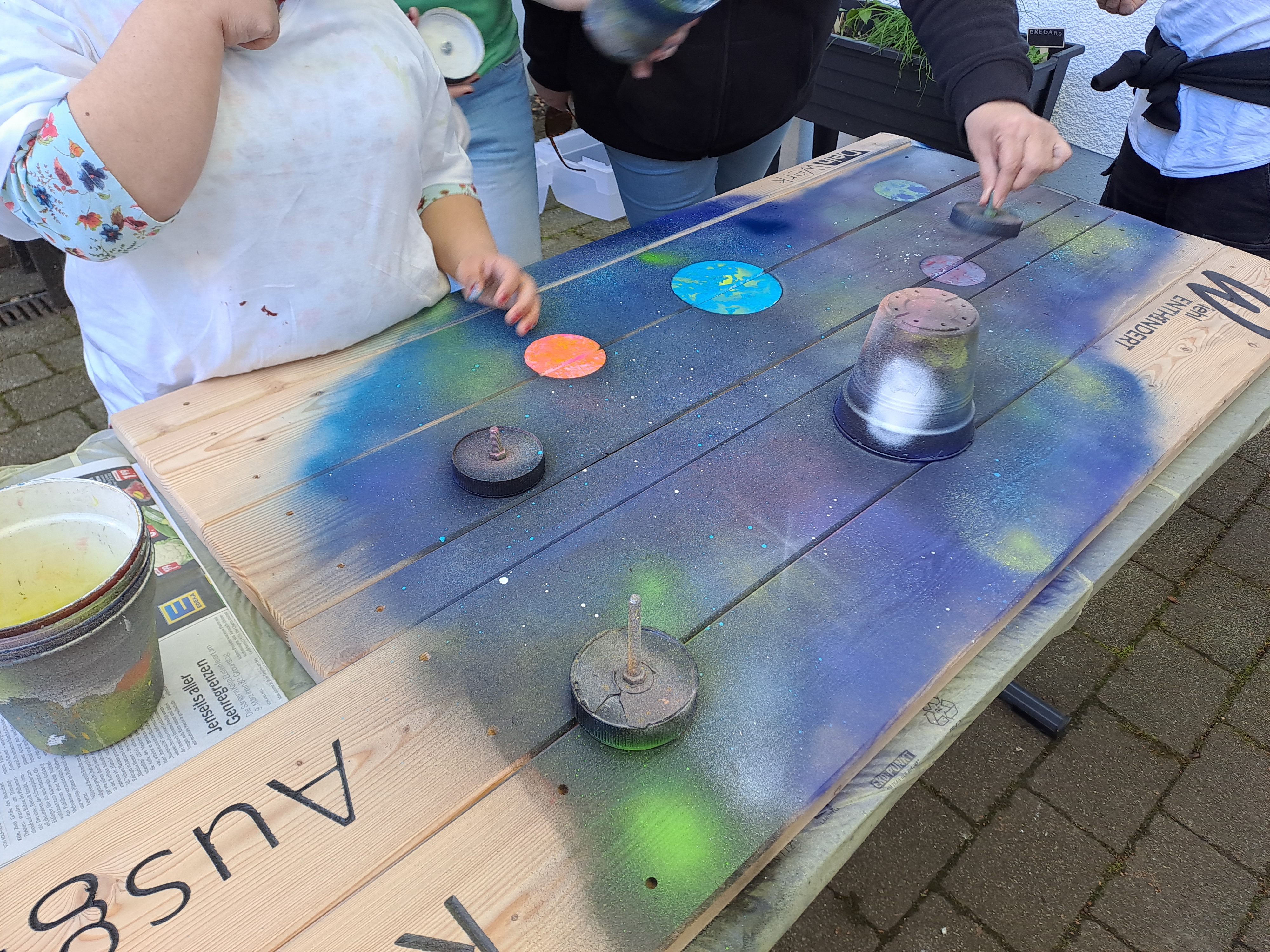 Man sprüht ein Weltraummotiv mit Planeten und Linien auf ein Holzbrett und verwendet Tassen und Deckel als Schablonen.