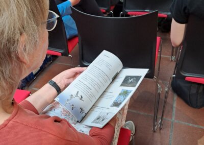 Eine Person, die in einer Stuhlreihe sitzt, liest während einer Veranstaltung in einer Halle ein Heft mit Vogelillustrationen.