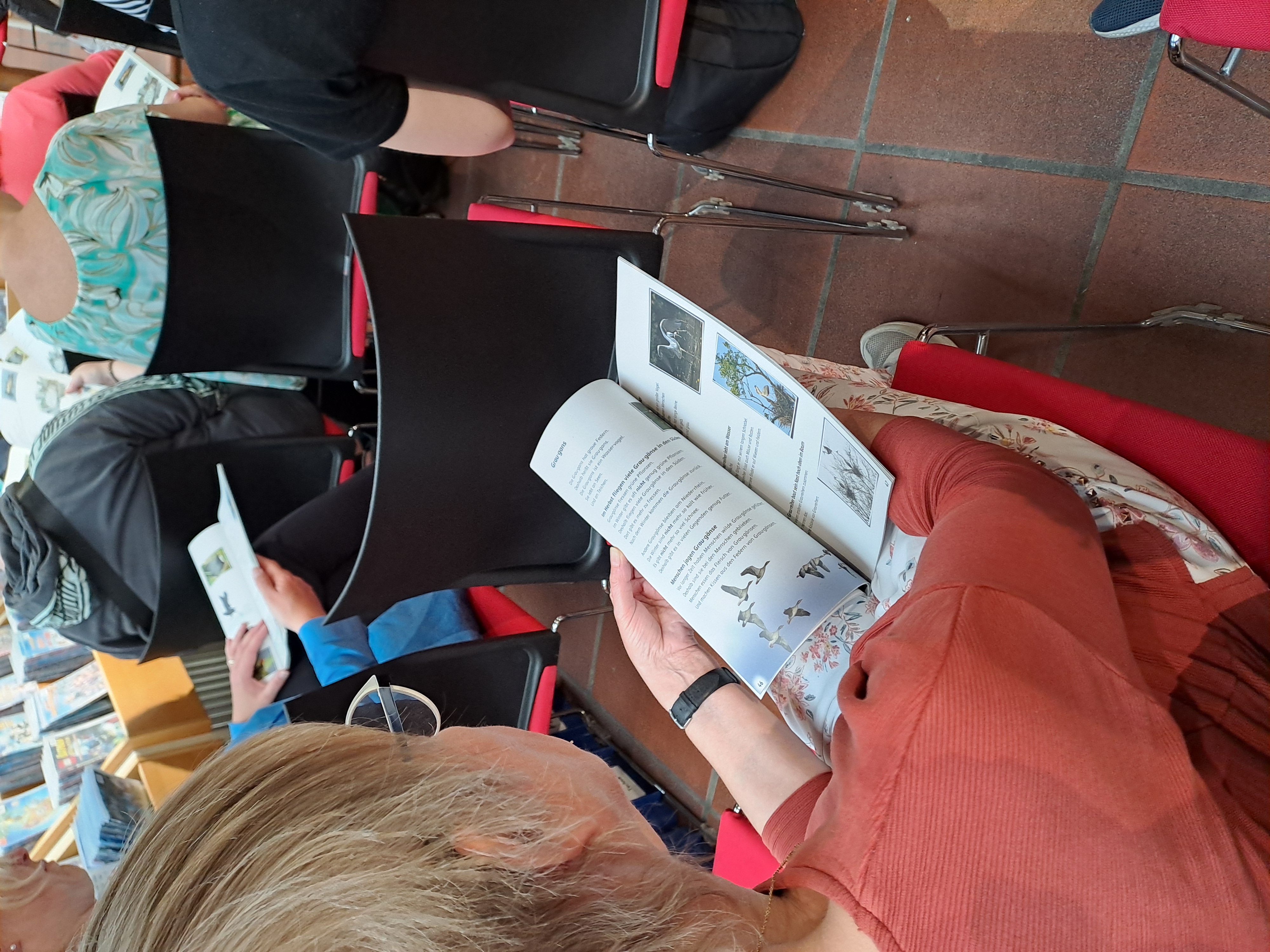Eine Person, die in einer Stuhlreihe sitzt, liest während einer Veranstaltung in einer Halle ein Heft mit Vogelillustrationen.