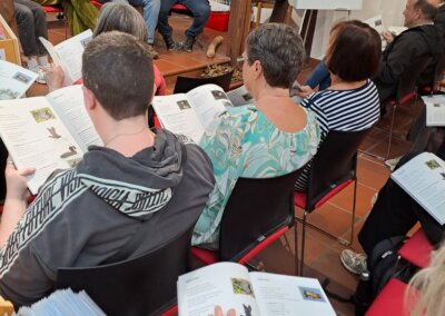 Eine Gruppe von Menschen sitzt in einer Bibliothek, liest Broschüren und hört einem Sprecher zu, der vorne neben einer Tafel mit einer Zeichnung sitzt.