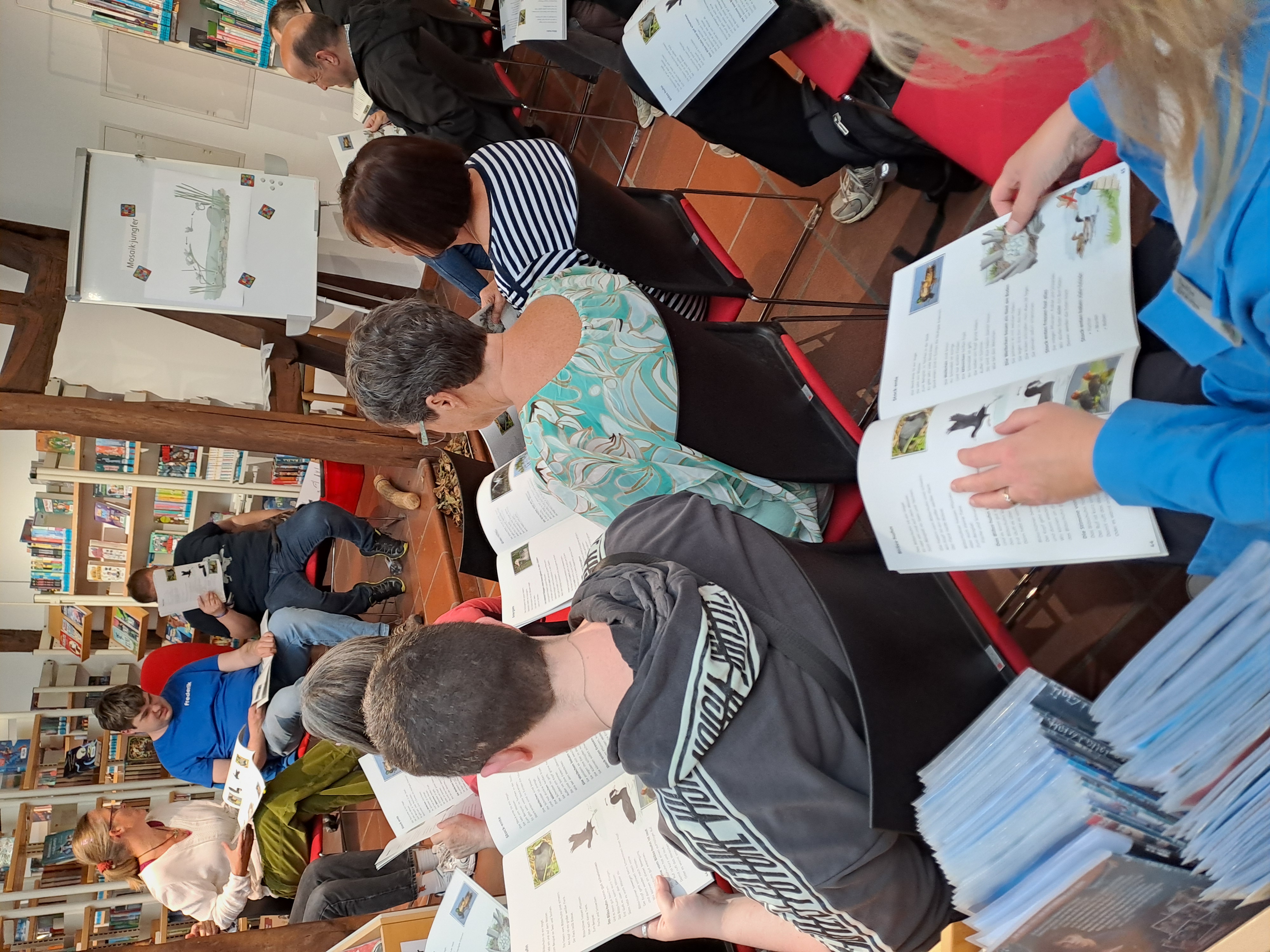 Eine Gruppe von Menschen sitzt in einer Bibliothek, liest Broschüren und hört einem Sprecher zu, der vorne neben einer Tafel mit einer Zeichnung sitzt.