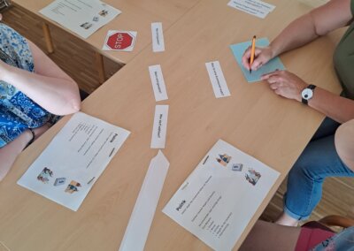 Drei Personen sitzen mit Dokumenten, Namensschildern und Notizkarten an einem Tisch und nehmen an einer Aktivität teil, bei der es darum geht, Papierstreifen mit aufgedruckten Wörtern anzuordnen.