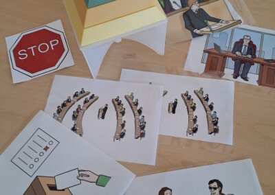 Eine Papierpyramide und verschiedene illustrierte Blätter zeigen auf einem Holztisch ein Stoppschild, eine Wahlurne, parlamentarische Szenen und Menschen im Gespräch.