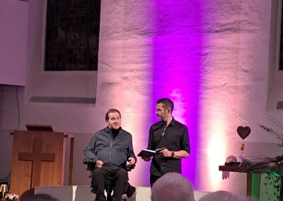 Zwei Männer stehen auf der Bühne vor einer beleuchteten Wand, der eine sitzt im Rollstuhl, der andere steht mit einer Tafel und spricht zu einem Publikum in einer Kirche.