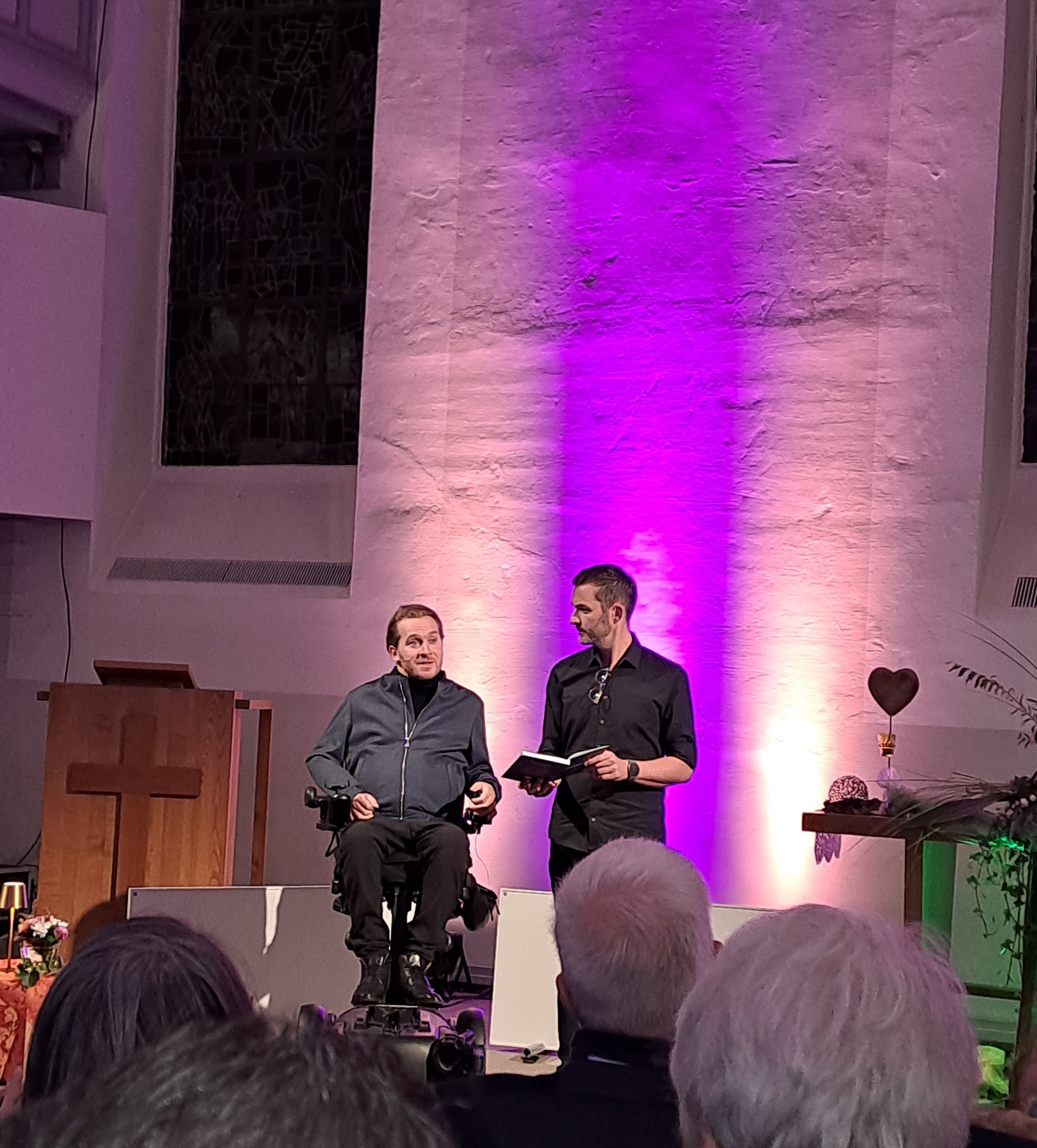 Zwei Männer stehen auf der Bühne vor einer beleuchteten Wand, der eine sitzt im Rollstuhl, der andere steht mit einer Tafel und spricht zu einem Publikum in einer Kirche.