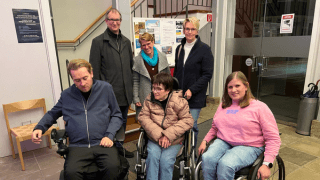 Sechs Erwachsene, darunter zwei in Rollstühlen, posieren gemeinsam in einem Innenraum vor einer Schautafel und einer Treppe.