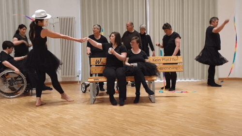 Eine Gruppe von schwarz gekleideten Menschen tritt in einem Raum auf; einige sitzen auf einer Bank mit der Aufschrift "Kein Platz für Ausgrenzung", während andere tanzen und mit Bändern hantieren.