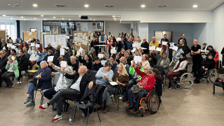 Eine große Gruppe von Menschen, darunter einige in Rollstühlen, sitzt in einem Raum, viele halten während einer Sitzung oder Veranstaltung Papiere hoch.