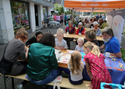 Eine Gruppe von Kindern und Erwachsenen sitzt an Tischen im Freien und beschäftigt sich unter einem Vordach bei einer Gemeindeveranstaltung mit Kunst und Handwerk.