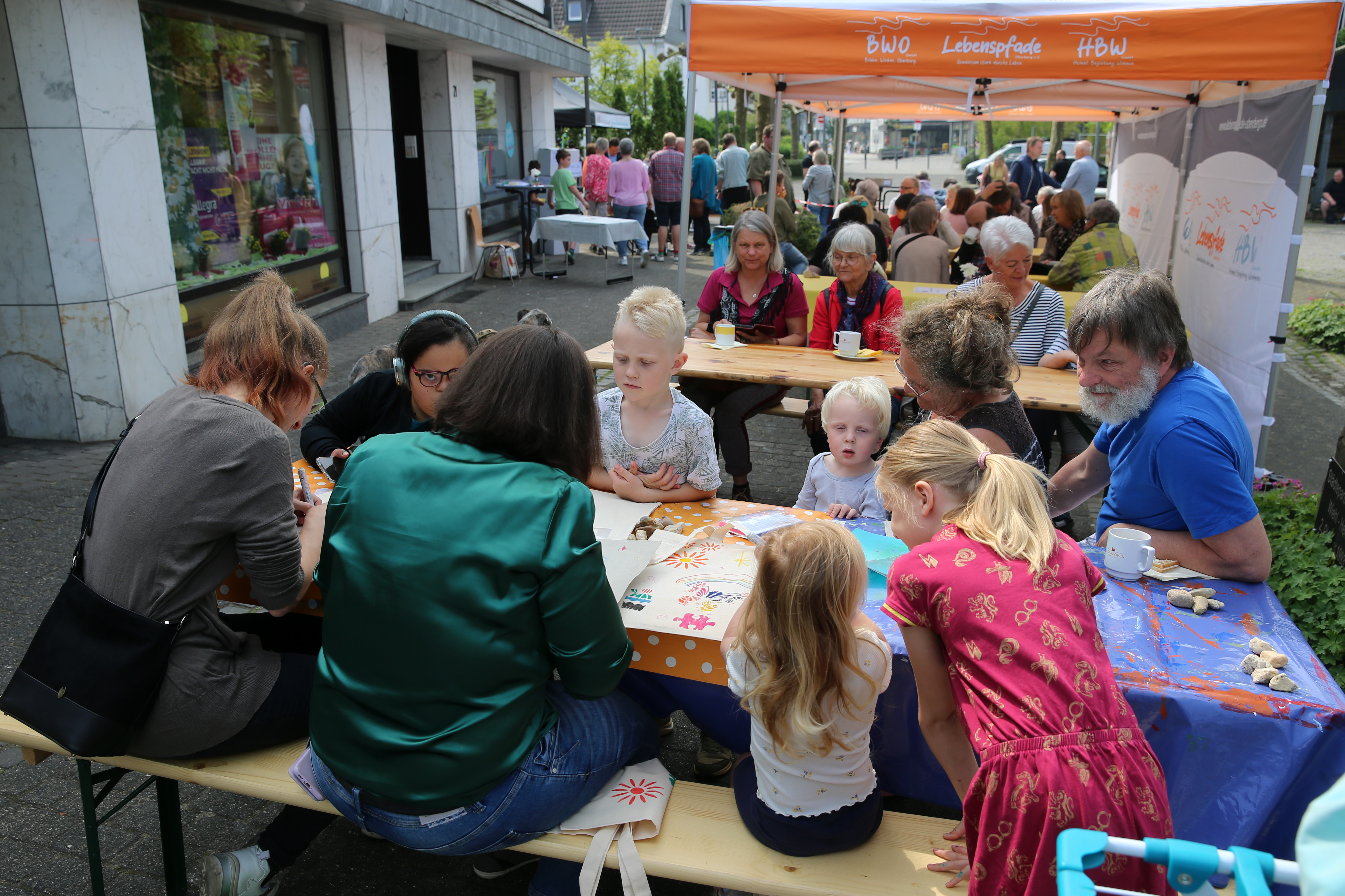 Eine Gruppe von Kindern und Erwachsenen sitzt an Tischen im Freien und beschäftigt sich unter einem Vordach bei einer Gemeindeveranstaltung mit Kunst und Handwerk.