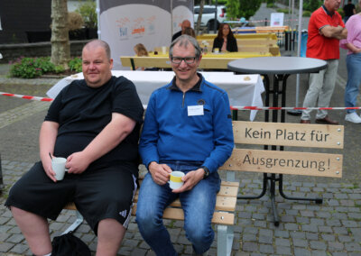Zwei Männer sitzen auf einer Bank mit einem Schild, auf dem "Kein Platz für Ausgrenzung! (Kein Platz für Ausgrenzung!) bei einer Veranstaltung im Freien und halten Tassen.