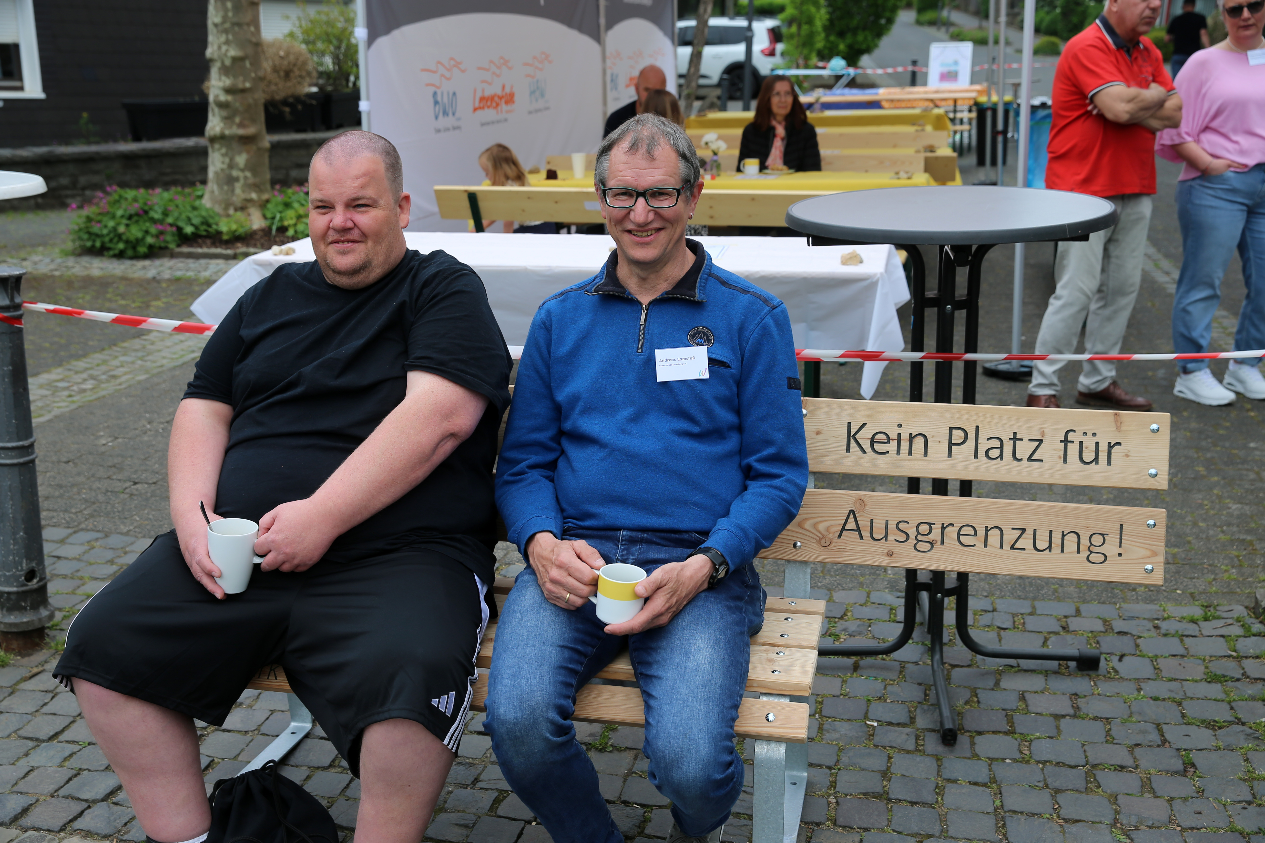 Zwei Männer sitzen auf einer Bank mit einem Schild, auf dem "Kein Platz für Ausgrenzung! (Kein Platz für Ausgrenzung!) bei einer Veranstaltung im Freien und halten Tassen.