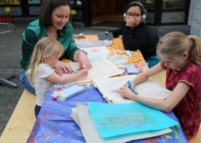 Drei Kinder und ein Erwachsener sitzen an einem Tisch im Freien und zeichnen und malen mit Markern und Schablonen auf Papier und Tragetaschen.