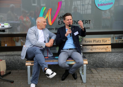 Zwei Männer in Anzügen sitzen auf einer Bank und unterhalten sich. Einer hält ein Mikrofon in der Hand. Hinter ihnen steht ein Schild mit der Aufschrift "Kein Platz für Ausgrenzung".