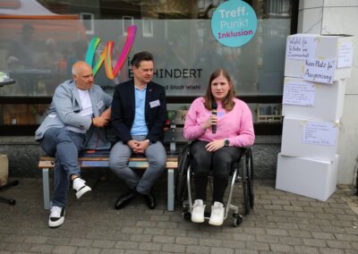 Drei Personen nehmen an einer Veranstaltung im Freien teil; zwei sitzen auf einer Bank, und eine Frau im Rollstuhl spricht in ein Mikrofon. In der Nähe sind Kisten mit Papieren gestapelt.