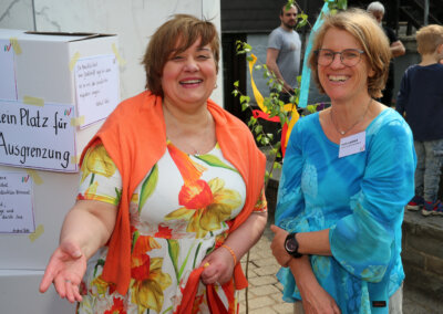 Zwei Frauen stehen und lächeln bei einer Veranstaltung im Freien; die eine trägt ein geblümtes Kleid mit einem orangefarbenen Schal, die andere ein blaues Oberteil und eine Brille. Im Hintergrund sind Menschen und Schilder mit deutschem Text zu sehen.
