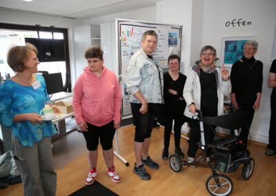Eine Gruppe von sechs Erwachsenen, einige mit sichtbaren Behinderungen, steht in einem hell erleuchteten Raum mit Kunstwerken und einem Flipchart im Hintergrund und interagiert.