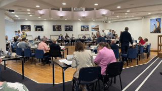 Eine Gruppe von Menschen sitzt und steht in einem großen Sitzungssaal um Tische herum und ist mit Diskussionen und Papierkram beschäftigt.