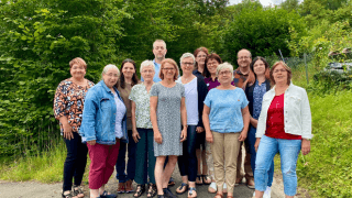 Eine Gruppe von vierzehn Erwachsenen, Männern und Frauen, steht im Freien auf einem Weg mit grünen Bäumen und Büschen im Hintergrund.