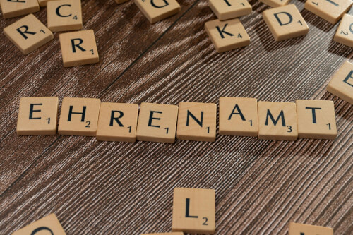 Auf einem Tisch liegen hölzerne Buchstabenplättchen, die das deutsche Wort EHRENAMT buchstabieren, umgeben von anderen verstreuten Buchstabenplättchen.