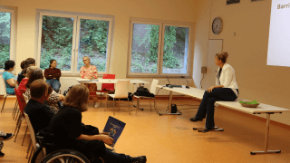 Eine Frau sitzt an einem Tisch im vorderen Teil eines Raumes und spricht zu einem sitzenden Publikum; eine Präsentation wird an die Wand projiziert.