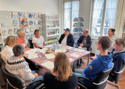 Neun Personen sitzen in einer Bibliothek oder einem Besprechungsraum um einen Tisch herum und diskutieren, auf dem Tisch liegen Papiere und Wasserflaschen, im Hintergrund stehen Bücherregale.