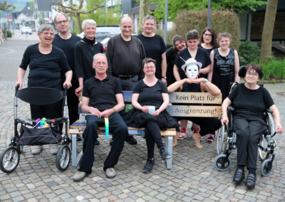 Eine Gruppe von Menschen, einige mit sichtbaren Behinderungen und Mobilitätshilfen, posiert im Freien mit einem Schild, auf dem "Kein Platz für Ausgrenzung" steht.