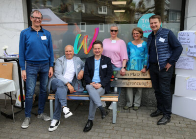 Sechs Erwachsene posieren und lächeln vor einem Gebäude, wobei zwei auf einer Bank sitzen und vier dahinter stehen. Sie tragen Namensschilder und Freizeit- oder Business-Casual-Kleidung. Es sind Schilder zum Thema Inklusion zu sehen.