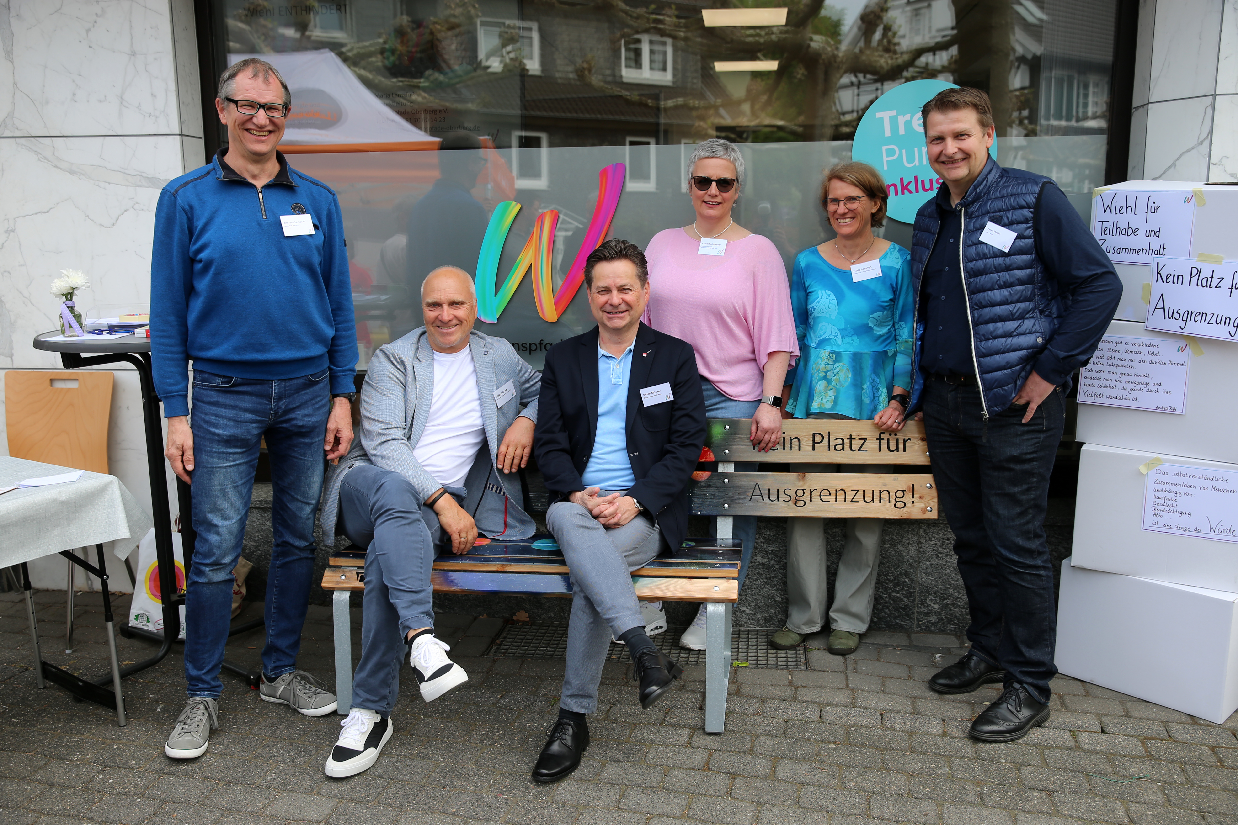 Sechs Erwachsene posieren und lächeln vor einem Gebäude, wobei zwei auf einer Bank sitzen und vier dahinter stehen. Sie tragen Namensschilder und Freizeit- oder Business-Casual-Kleidung. Es sind Schilder zum Thema Inklusion zu sehen.