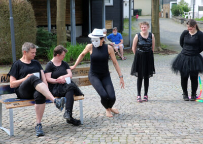 Fünf Personen befinden sich im Freien; eine Person mit einer weißen Maske und einem Hut posiert vor zwei sitzenden Personen, während zwei weitere Personen in schwarzen Kleidern in der Nähe stehen.
