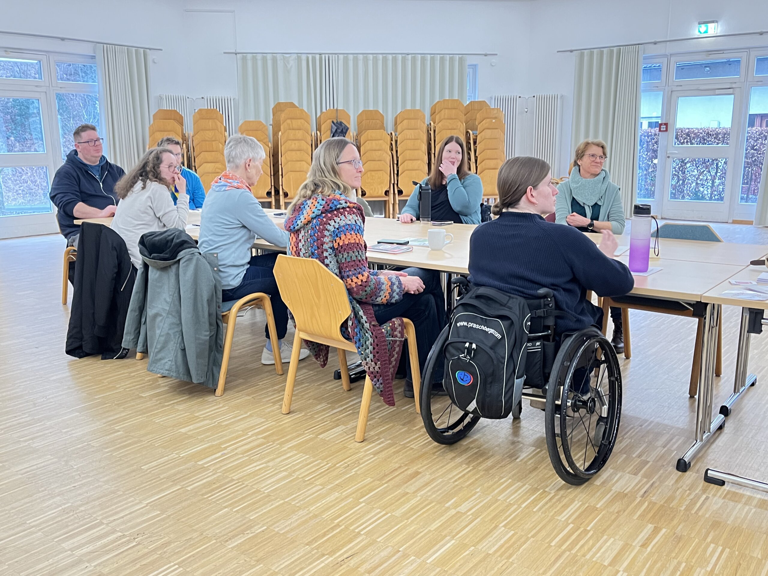 9 personen sitzen um einen tisch und hören einer sprecherin zu